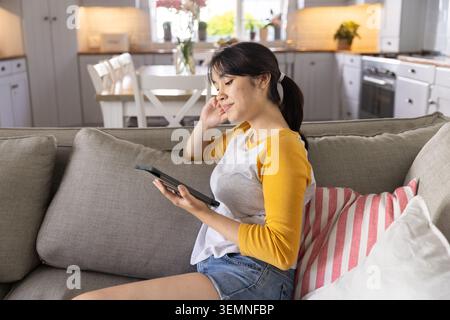 Asiatische Frau, die auf Sofa mit Kissen im offenen Wohnbereich sitzt und ein Tablet hält Stockfoto