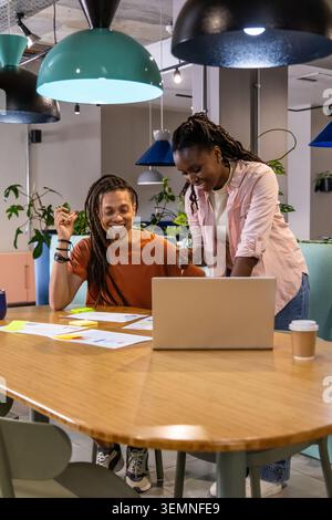 Afroamerikanische Kollegen sitzen und lehnen sich am Tisch im Büro und zeigen auf Laptop und Dokumente Stockfoto
