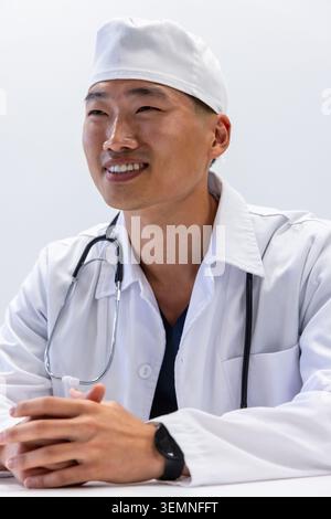 Asiatischer Arzt sitzt in der Klinik, Hände auf weißem Tisch, trägt Labormantel, Mütze und Stethoskop Stockfoto