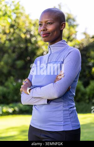Afroamerikanerin, die im Park steht, mit Viertelreißverschluss, Leggings, überkreuzten Armen, Smartwatch. Fitness, Outdoor, sportlich, selbstbewusst, Sonnenlicht, Natu Stockfoto