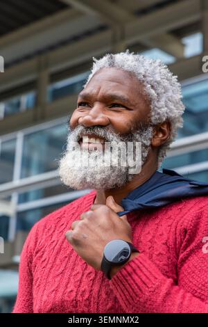 Lächelnder Mann mit weißem Bart, der Jacke an der modernen Fassade hält, roter Pullover und Smartwatch trägt Stockfoto
