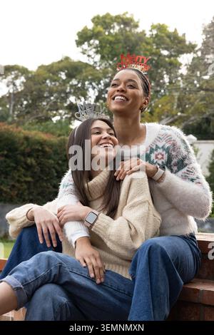 Verschiedene weibliche Freunde sitzen auf Ziegelwänden im Garten und tragen Strickpullover Stirnbänder Smartwatches Stockfoto