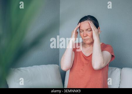 kaukasische Frau mittleren Alters, die unter starken Kopfschmerzen leidet, während sie auf einem Balkonsofa sitzt. Selektiver Fokus. Stockfoto