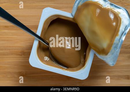 Karamell Puddingbecher mit Löffel auf hölzernem Hintergrund, Nahaufnahme. Eine offene Plastiktasse mit glattem Karamell oder Kaffeepudding Dessert mit einem Metalllöffel Insis Stockfoto