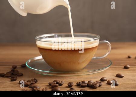 Milch in Glasbecher und Bohnen auf Holztisch gießen, Nahaufnahme Stockfoto