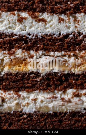 Nahaufnahme des Texturhintergrunds eines geschichteten Schokoladenkuchens mit Sahnefüllung. Makroaufnahme aus Schokoladenschwamm-Kuchenschichten mit weißer Creme und Bananenkaramat Stockfoto