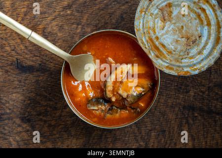 Blick von oben auf geöffnete Dosenfischsprossen in roter Tomatensoße mit einer goldenen Gabel auf einem dunklen Holztisch, Nahaufnahme Stockfoto