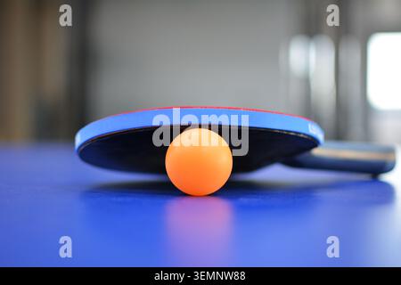 Nahaufnahme eines orangefarbenen Tischtennisballs und Tischtennispaddels auf einem blauen Tisch, mit verschwommenem Bild, das eine konzentrierte, aber dynamische Sportszene erzeugt. Stockfoto