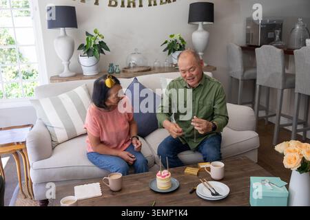 Asiatisches Paar, reif und älter, sitzt zu Hause auf dem Sofa und legt Kerzen auf Kuchen mit Geschenk Stockfoto