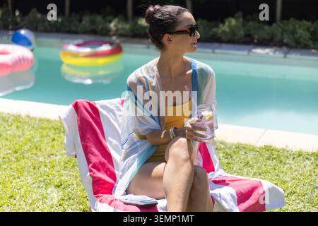 Frau mittleren Alters, die auf einem mit Handtüchern überzogenen Liegestuhl am heimischen Pool sitzt, in gelben Badeanzügen mit einem Glas Stockfoto