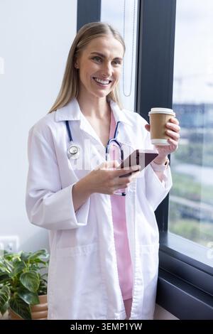Weibliche Ärztin, die in der Klinik steht und Laborkittel trägt, rosa Peelings, Stethoskop, das Telefon und Kaffee hält Stockfoto