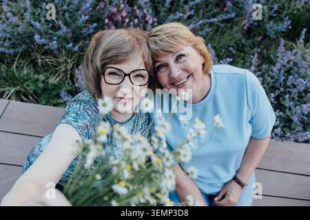 Lächelnde Seniorin und Erwachsene Tochter sitzen zusammen in der Nähe von Wildblumen Stockfoto