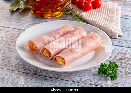 Drei Scheiben Schinken gerollt mit herzhafter Füllung auf weißem Teller. Frische Petersilie, Kirschtomaten, Rosmarin, infundiertes Öl auf rustikalem Holztisch Stockfoto