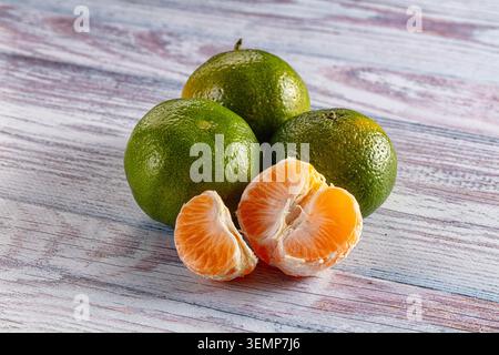 Drei unreife grüne Mandarinen mit strukturierter Haut sitzen auf verwitterter Holzoberfläche neben geschälten Halbfrüchten, die saftig orange wirken Stockfoto