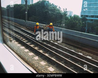 Zwei männliche Eisenbahner, die orangefarbene Sicherheitswesten und gelbe Schutzhelme tragen, heben Ausrüstung auf einer Eisenbahnstrecke an Stockfoto