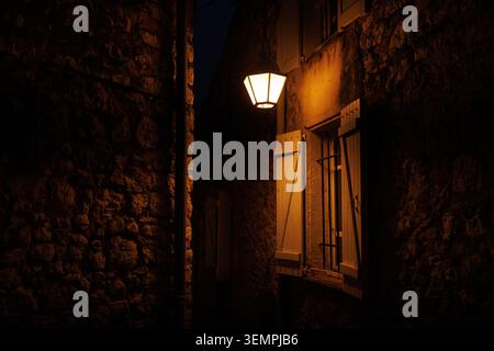 Eine dramatische Aufnahme einer glühenden Laterne, die ein warmes Licht auf eine Hausfassade aus Stein in einer dunklen Gasse wirft. Stockfoto