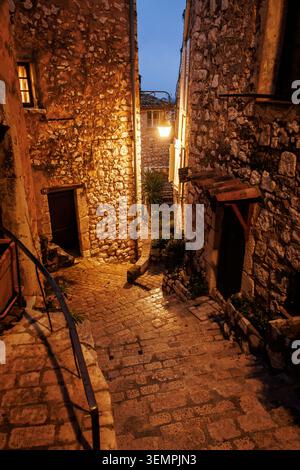 Ein stimmungsvoller Blick auf eine leere Kopfsteinpflasterallee, die sich zwischen rustikalen Steinmauergebäuden schlängelt, die von einer goldenen Straßenlaterne beleuchtet werden. Stockfoto