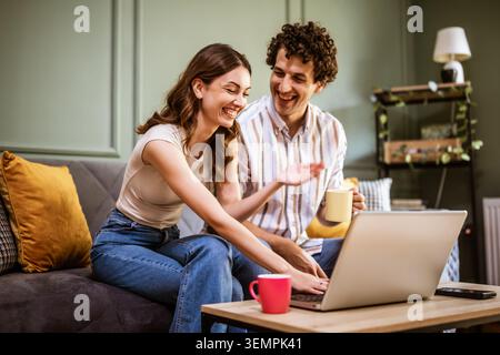Zwei junge Erwachsene sitzen eng auf einer Couch, konzentrieren sich auf einen Laptop und teilen einen angenehmen Moment. Stockfoto