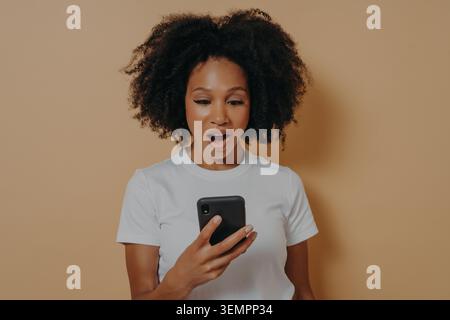 Überraschte junge afrikanische Frau, die mit schockiertem Gesichtsausdruck auf das Smartphone schaut, unerwartete Nachrichten im Internet liest oder mit einem Freund chatet, in weißem T-Shirt gekleidet, isoliert über beigefarbenem Hintergrund Stockfoto