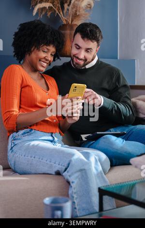 Vertikale Aufnahme fröhlicher junger Mann und Frau, die auf einem bequemen Sofa sitzen, lächeln und zusammen auf einen Smartphone-Bildschirm schauen, zu Hause, im Lifestyle, in der Freizeit Stockfoto