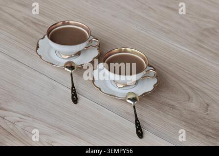 Zwei Tassen Kaffee mit Milch in antiken Kaffeetassen mit silbergoldenen Löffeln auf Untertassen auf einem Eichentisch. Nahaufnahme der Vorderansicht. Stockfoto