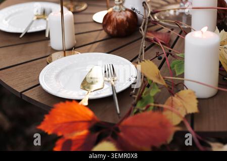 Eleganter Herbsttisch im Freien mit weißen Tellern, goldenem Besteck, beleuchteten Kerzen und Kürbissen inmitten farbenfroher Herbstblätter für ein gemütliches Erntedinn Stockfoto