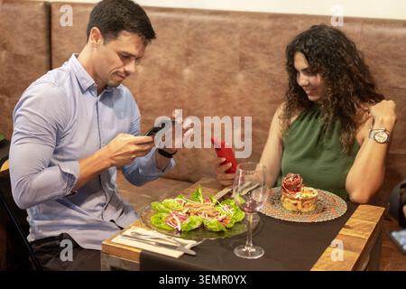 Junge Männer und Frauen, die ihre Smartphones benutzen, während sie zusammen in einem Restaurant essen. Moderner Lebensstil, Mobiltechnologie, digitale Kommunikation, soziale Medien Stockfoto