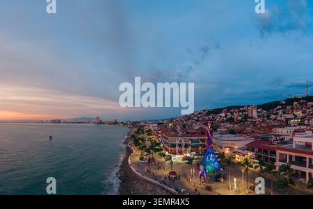 Blick aus der Vogelperspektive auf die beleuchtete Promenade von Malecon mit Menschen, die an einer riesigen Catrina-Statue und Küstengebäuden in der Abenddämmerung in Puerto Vallarta, JAL, vorbeilaufen Stockfoto
