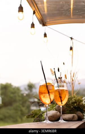 Auf einem rustikalen Holztisch stehen zwei elegante Stielgläser mit leuchtendem Orangencocktail, Eiswürfeln und schwarzen Strohhalmen Stockfoto