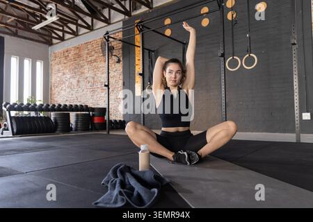 Erwachsene Frau, die sich auf Matte im Lagerhaus-Fitnessstudio dehnt, trägt schwarze Sportbekleidung mit Wasserflasche Stockfoto