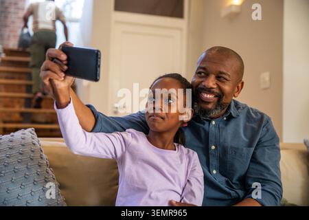 Afroamerikanischer Vater und Kind machen Selfie auf beigefarbener Couch, Kind hält Smartphone Stockfoto