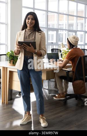Stehende Frau tippt auf dem Tablet und zeigt Geschäftsdiagramme auf dem gemeinsam genutzten Schreibtisch-Laptop zur Überprüfung an Stockfoto