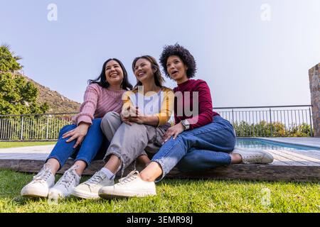 Verschiedene weibliche Freunde sitzen auf hölzernen Terrassenrändern am Pool auf grasbewachsenem Rasen und tragen weiße Sneakers Stockfoto