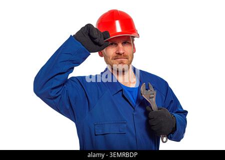 Arbeiter in blauem Overall und rotem Schutzhelm hält einen verstellbaren Schraubenschlüssel vor weißem Hintergrund. Stockfoto