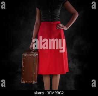 Studio-Aufnahme einer Frau in Vintage-Kleidung, die einen braunen Koffer vor dunklem Hintergrund hält. Stockfoto