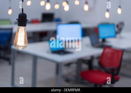 Eine Nahaufnahme mit selektivem Fokus einer glühenden Vintage-Glühlampe, die von der Decke hängt, mit einem verschwommenen, leeren modernen Technikbüro, Schreibtisch Stockfoto