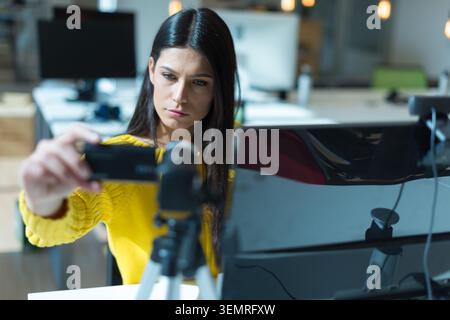 Eine fokussierte junge Geschäftsfrau in einem gelben Pullover greift nach vorne, um eine kleine Videokamera auf einem Stativ auf ihrem Schreibtisch einzustellen. Stockfoto