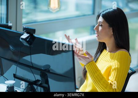 Hochwinkelansicht einer jungen Frau mit langen dunklen Haaren, die einen gelben Strickpullover trägt, während eines Online-Meetings mit beiden Händen spricht und Gesten macht Stockfoto