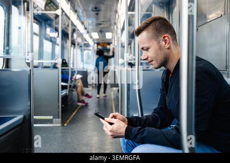Passagiere, die im Pendlerzug sitzen und während der täglichen Fahrt auf den Bildschirm des Mobiltelefons schauen. Öffentliche Verkehrsmittel, städtischer Lifestyle, Smartphone-Technologie und DIG Stockfoto