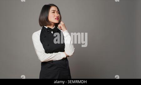 Die Gastgeberin des Restaurants ist aufmerksam über grauen Hintergrund und denkt über neue Ideen nach, um das Menü für Gäste zu entwickeln, mit asiatischer Kellnerin, die nachdenklich ist, Fantasie im Studio. Stockfoto