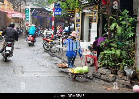 Ein Straßenverkäufer verwendet eine traditionelle Schulterstütze, um Produkte auf einer Straße in Hanoi, Vietnam, an Motorrädern vorbei zu transportieren Stockfoto