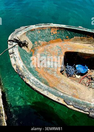 Ein altes Boot ist an einem Dock in Sliema, Malta, gebunden. Das Sonnenlicht reflektiert das grüne Wasser in der Nähe. Der Bereich weist Anzeichen von Verschleiß und Alter auf. Stockfoto