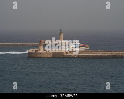 Eine Fähre nähert sich dem Hafen in Birgu, Malta, mit Leuchttürmen, die am Pier unter bewölktem Himmel sichtbar sind. Stockfoto