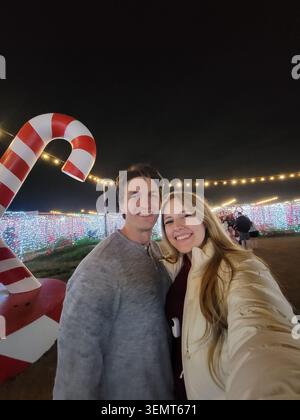 Ein junger Junge lächelt und posiert nahe beieinander bei einer nächtlichen Weihnachtsbeleuchtung im Freien neben einer großen rot-weiß gestreiften Zuckerrohr-Requisite. Warmes LED Stockfoto