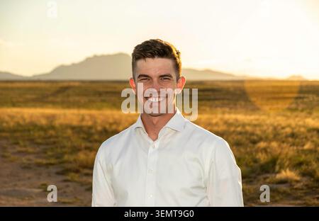 Ein fröhlicher Mann mit weißem Hemd steht während der goldenen Stunde in einer offenen Wüstenlandschaft. Die Szene zeigt entfernte Hügel und eine Gebirgskette, Krieg Stockfoto