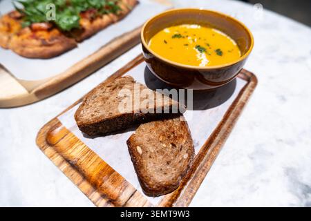 Warme Kürbissuppe serviert mit rustikalem Vollkornbrot auf Holzbrett, garniert mit frischen Kräutern, Stockfoto