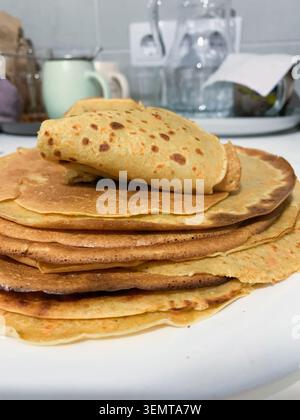 Pfannkuchen werden ordentlich auf einem weißen Teller in einer Küche gestapelt. Der Hintergrund zeigt verschiedene Küchenartikel wie Tassen und Gläser. Die Szene sieht warm aus Stockfoto