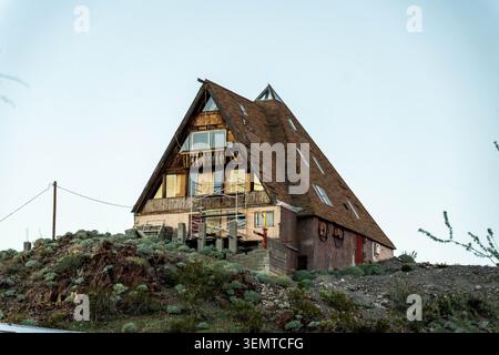 Ein Holzhaus mit A-Rahmen befindet sich auf einem felsigen Hügel mit einem steil geneigten Dach, großen Fenstern und einem rauen Äußeren. Die Struktur erhebt sich über verstreute s Stockfoto