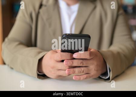 Ein Nahaufnahme konzentriert sich auf die Hände eines mittelgroßen asiatischen Mannes, der sein Handy über einem Tisch in einem Waschsalon mit geringer Schärfentiefe hält. Stockfoto