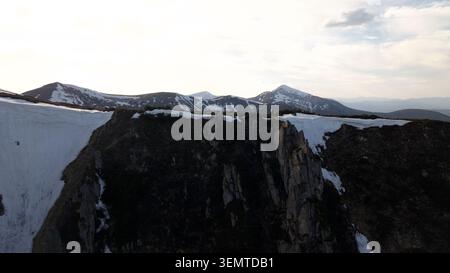 Drohnenperspektive von zwei winzigen Figuren, die auf einem dramatischen felsigen Kamm mit dicken Schneegesimsen vor dem Hintergrund majestätischer Berggipfel stehen Stockfoto